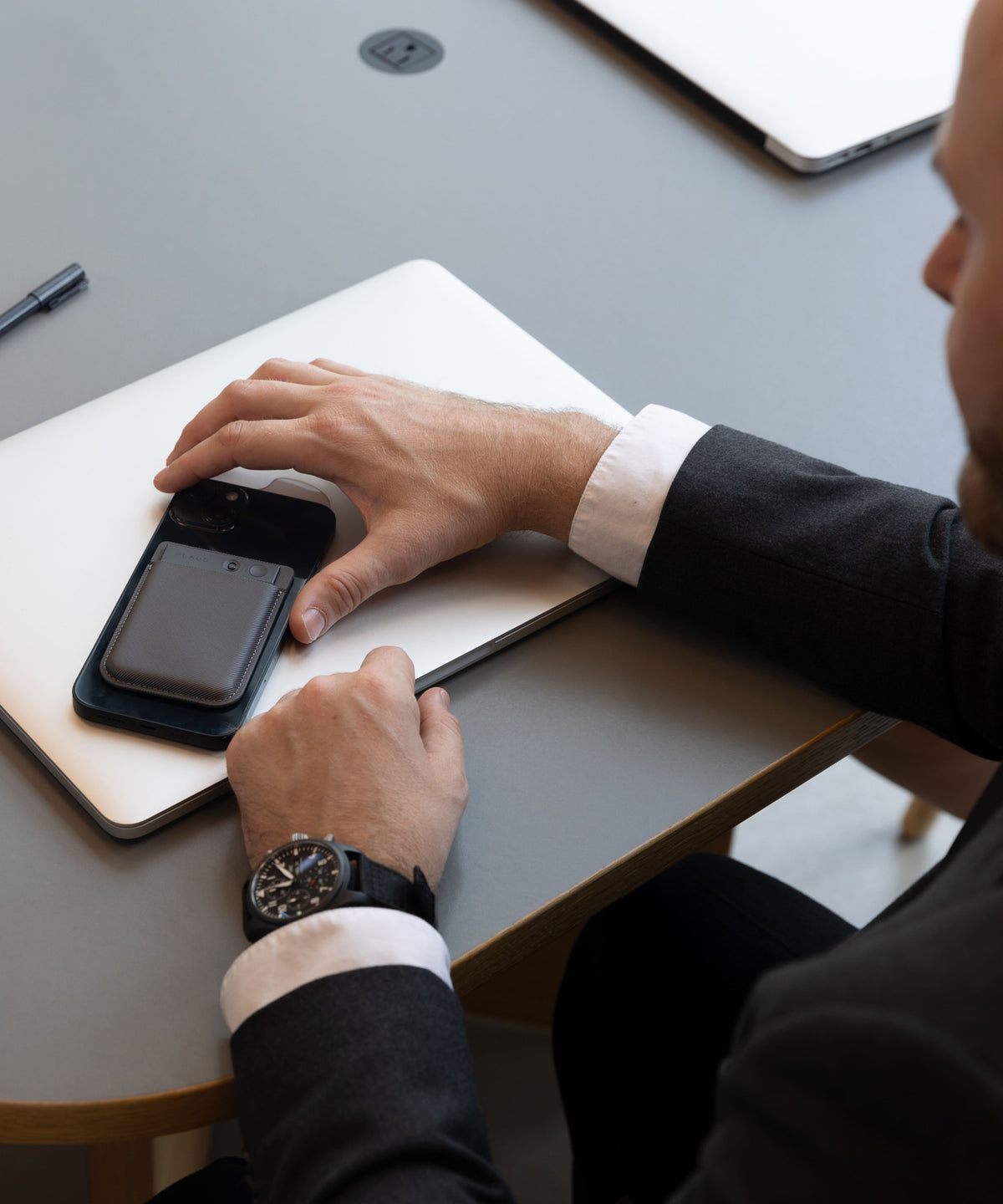 A professional in a suit uses a PLAUD NOTE AI voice recorder attached to a smartphone on a desk with a laptop.