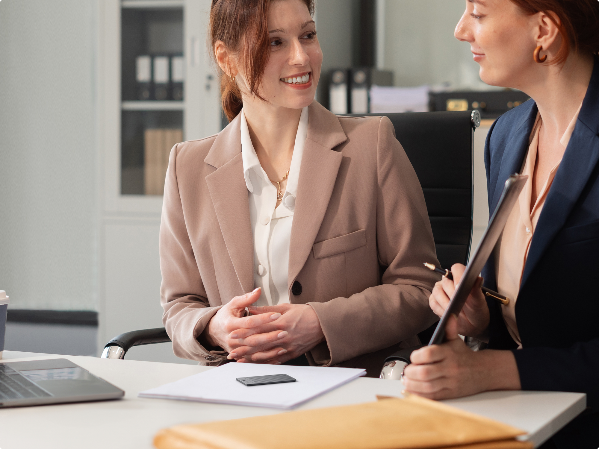 Two professionals collaborating in an office with a PLAUD NOTE on the desk and a PLAUD NotePin worn on a collar.