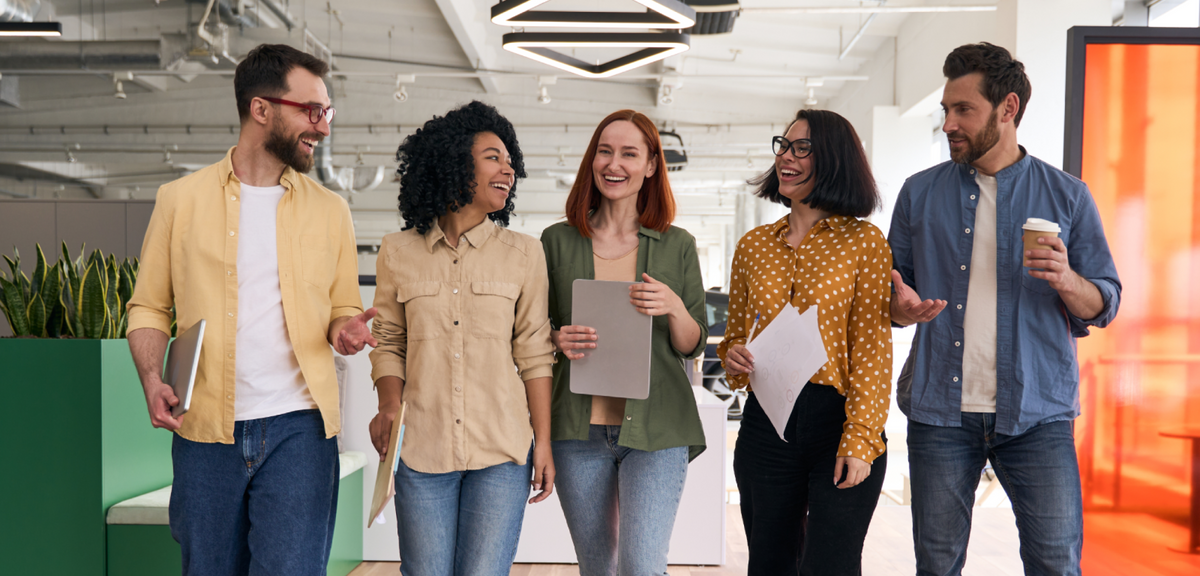 Five diverse professionals smiling and talking while walking through a modern, open-plan office.