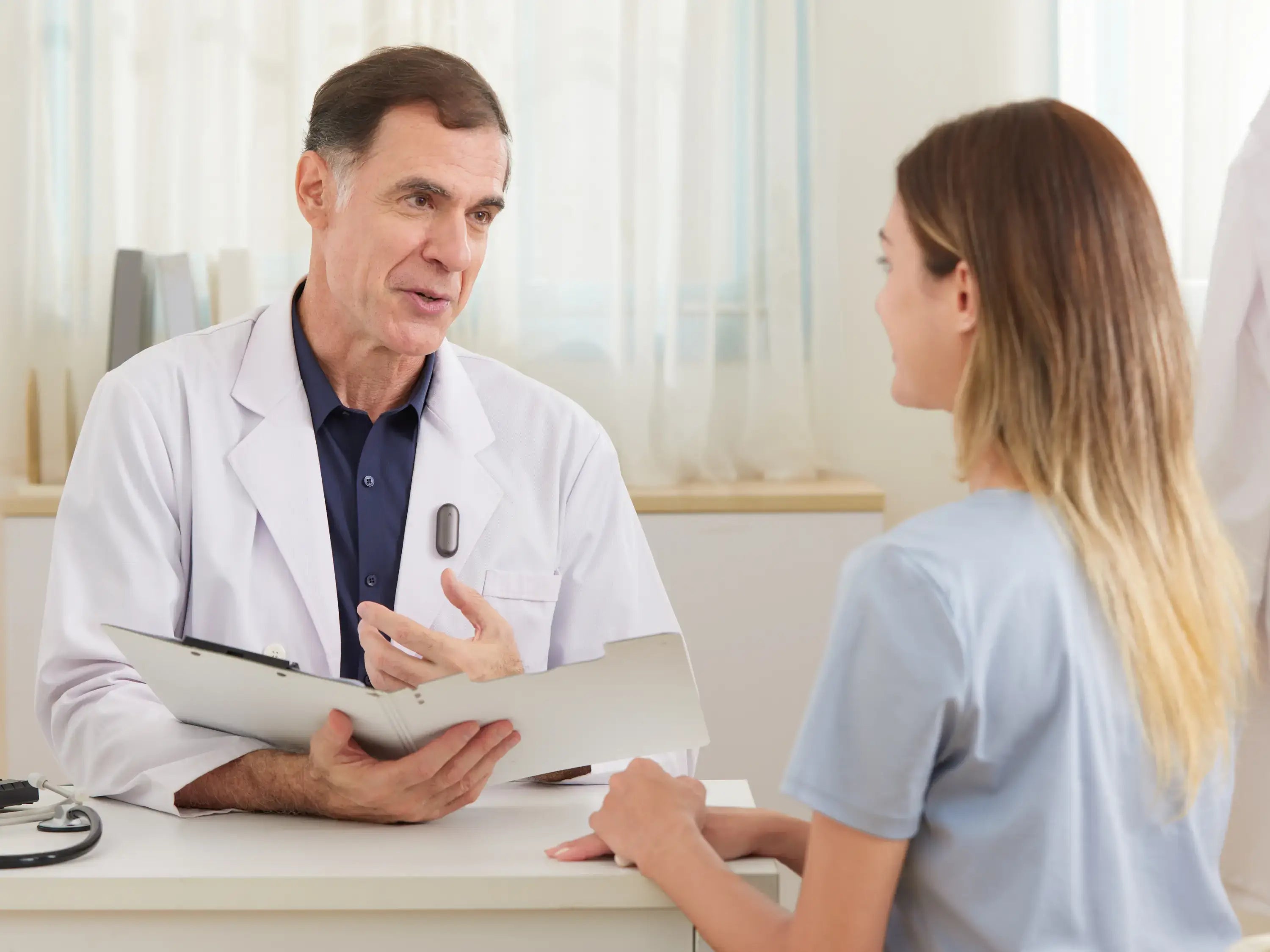 A doctor wearing a Plaud NotePin on his lapel while consulting with a patient in a medical office.