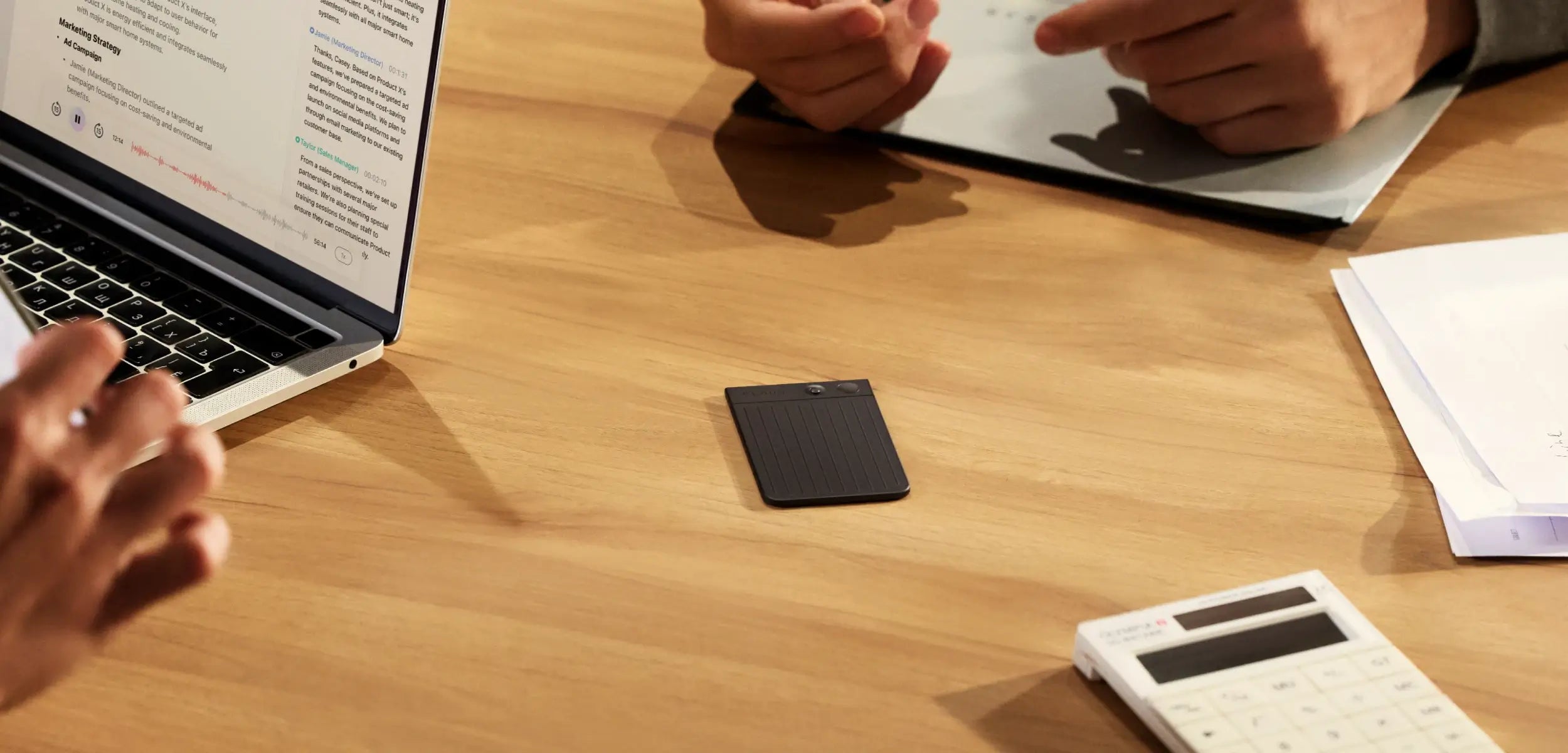 A black PLAUD NOTE voice recorder sits on a wooden table during a meeting with a laptop and calculator.
