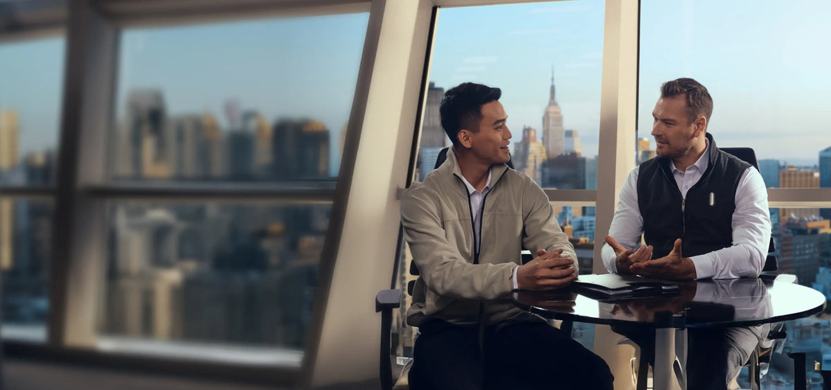 Two professionals in a high-rise office meeting; one man wears a Plaud NotePin to capture the conversation.