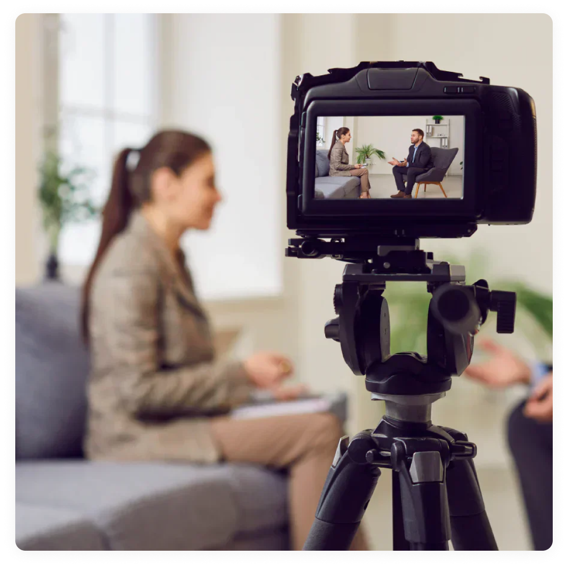 A camera on a tripod records a seated interview between a woman and a man in a bright office.