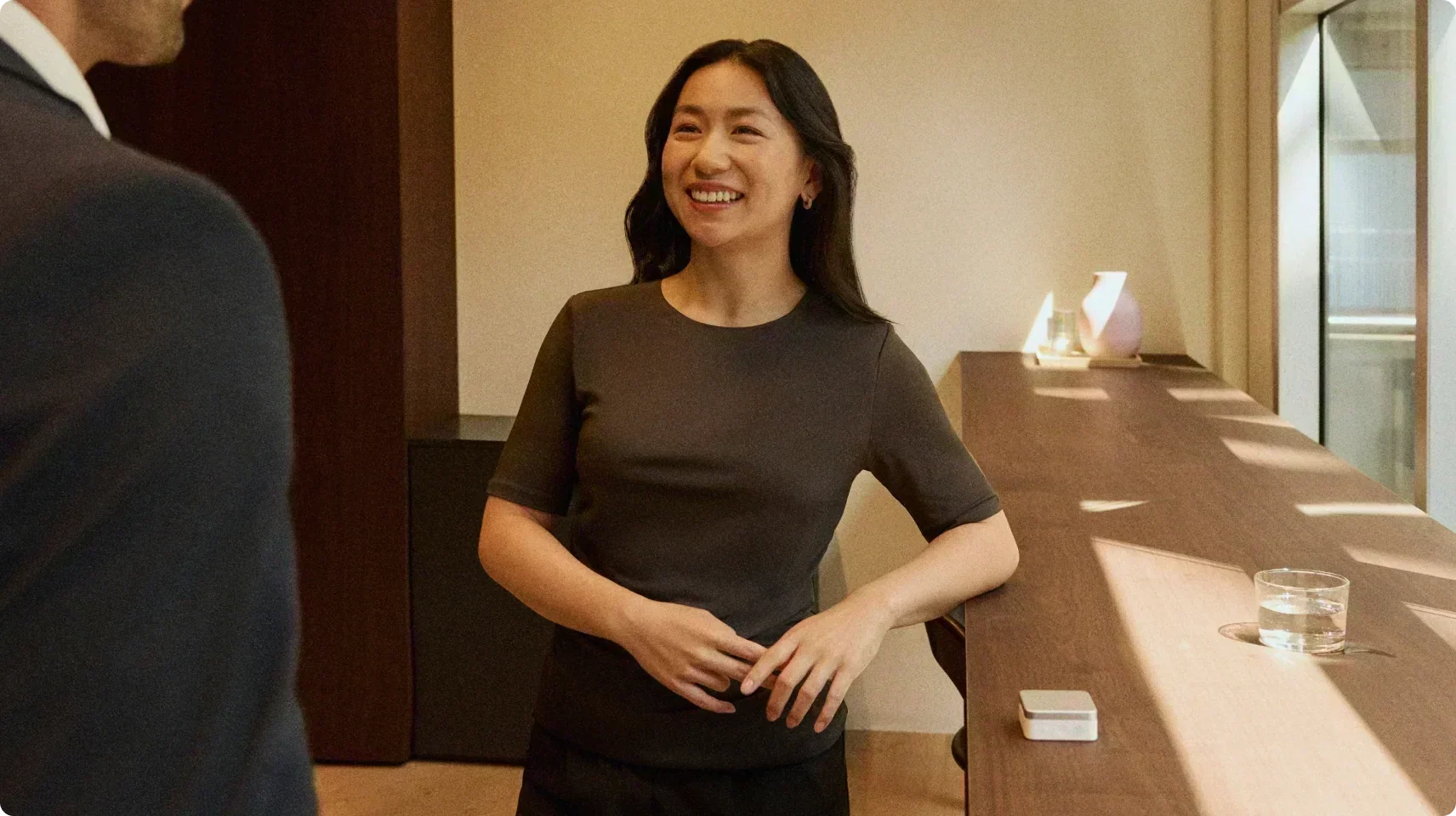 A woman smiles while talking to a colleague, with a silver Plaud Note recording the conversation from a wooden table.