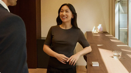 A woman smiles while talking to a colleague, with a silver Plaud Note recording the conversation from a wooden table.