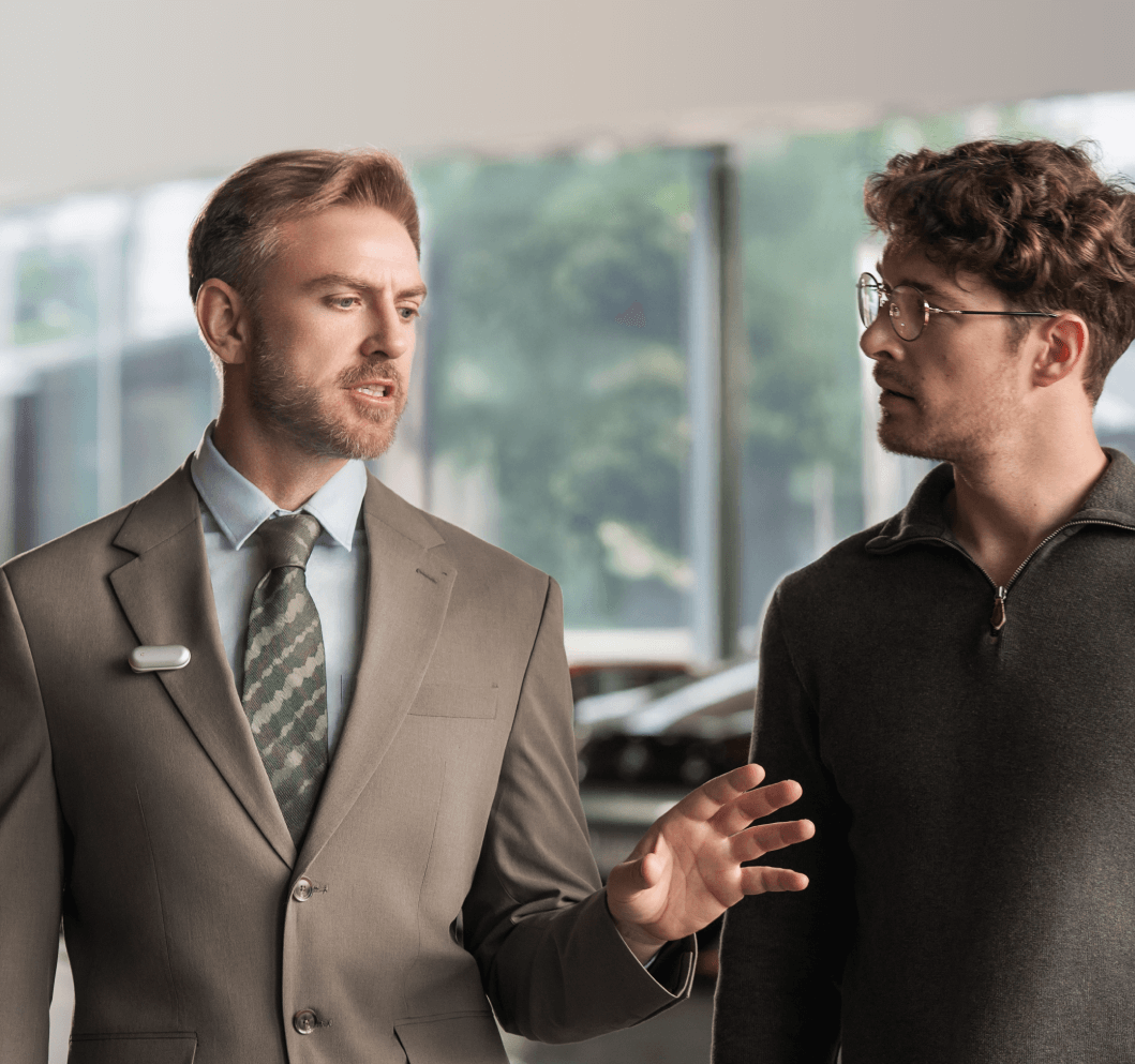A man in a suit wearing the Plaud NotePin on his lapel while speaking with a colleague.