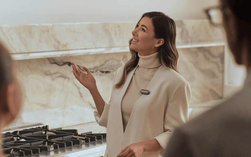 A professional woman wearing a Plaud NotePin on her lapel while speaking to clients in a modern kitchen.