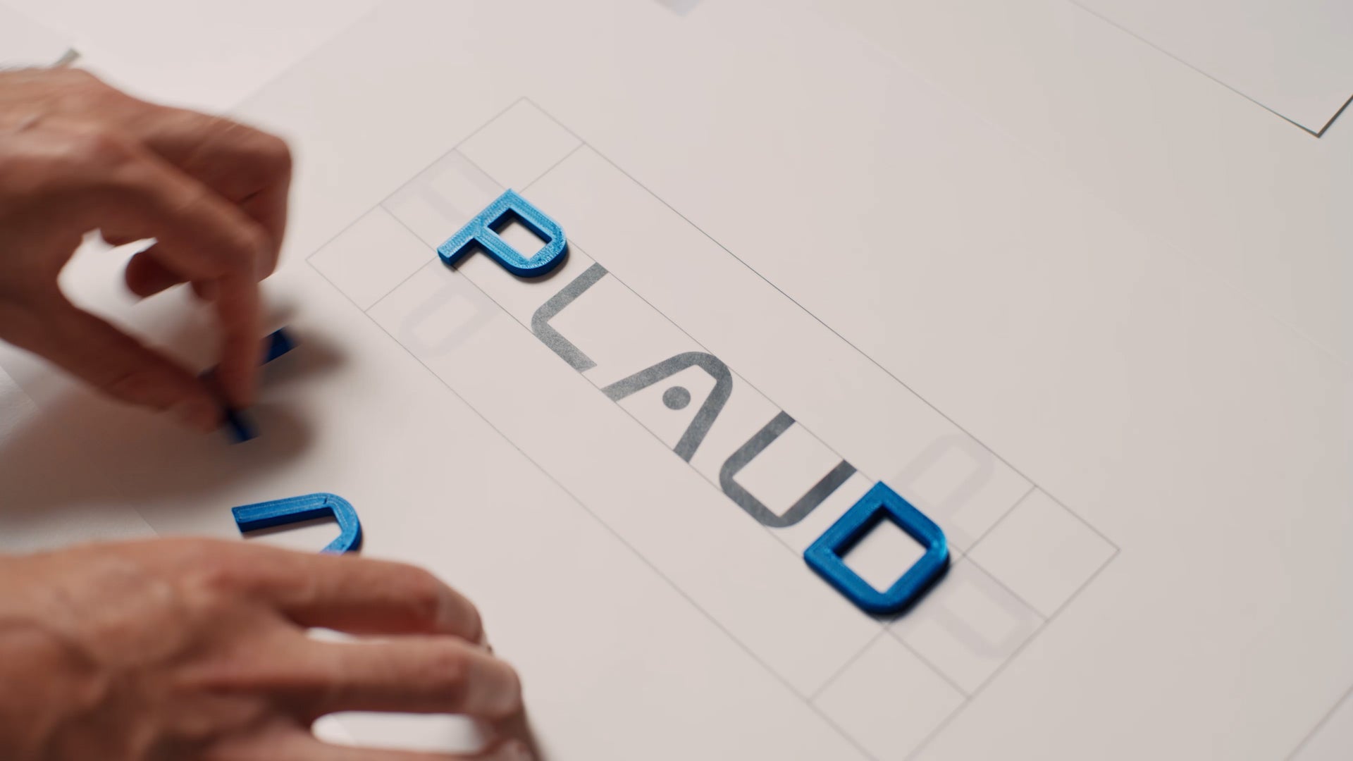 Hands arranging blue 3D letters to spell out the PLAUD brand name on a design grid.