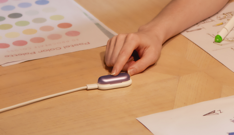 A hand presses the button on a purple Plaud NotePin in its charging dock on a desk with art supplies.
