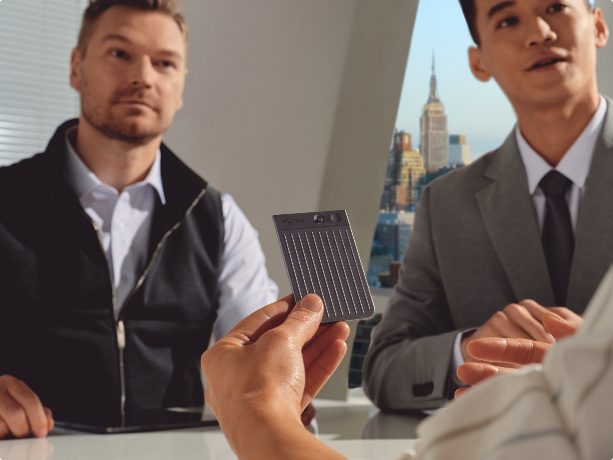 A person holds a Plaud Note AI voice recorder during a business meeting in an office with a city view.