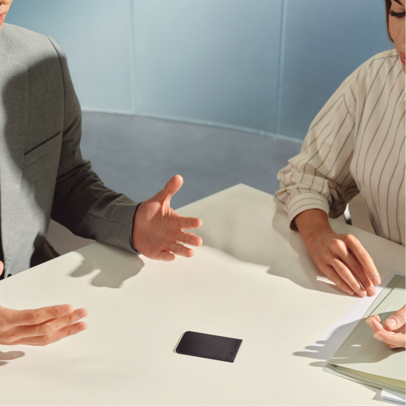 A Plaud Note AI voice recorder sits on a table between two people during a business meeting.
