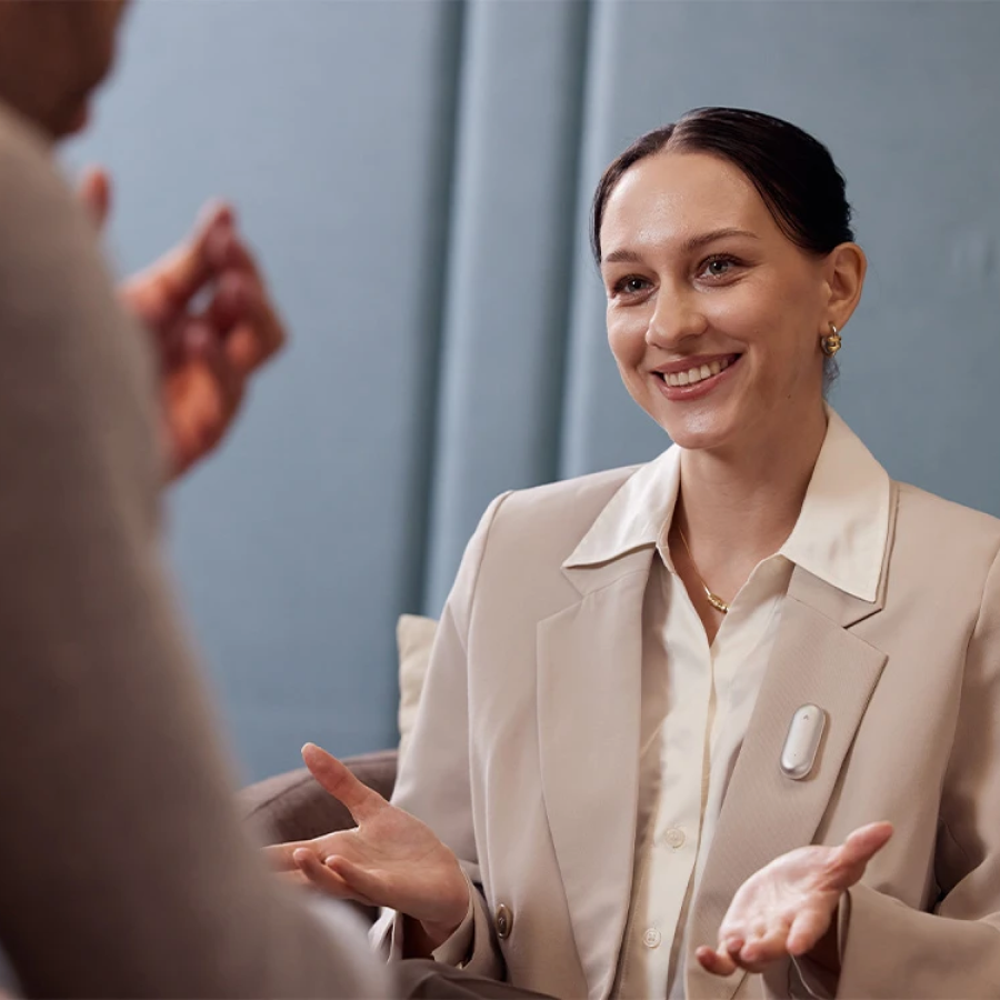 A smiling woman wearing the Plaud NotePin AI voice recorder on her blazer during a professional meeting.