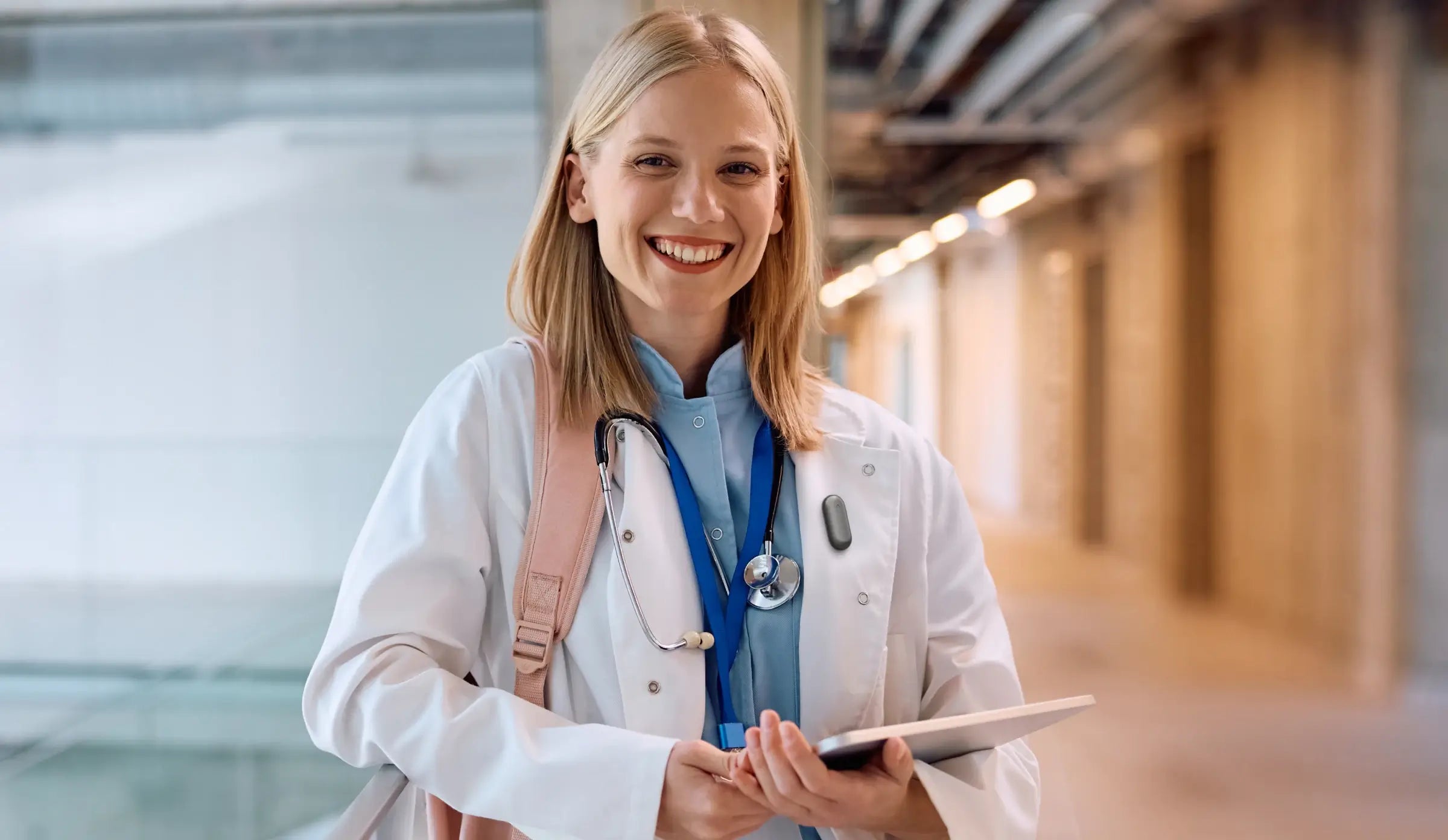 Smiling healthcare professional wearing a Plaud NotePin on her lab coat lapel in a hospital hallway.