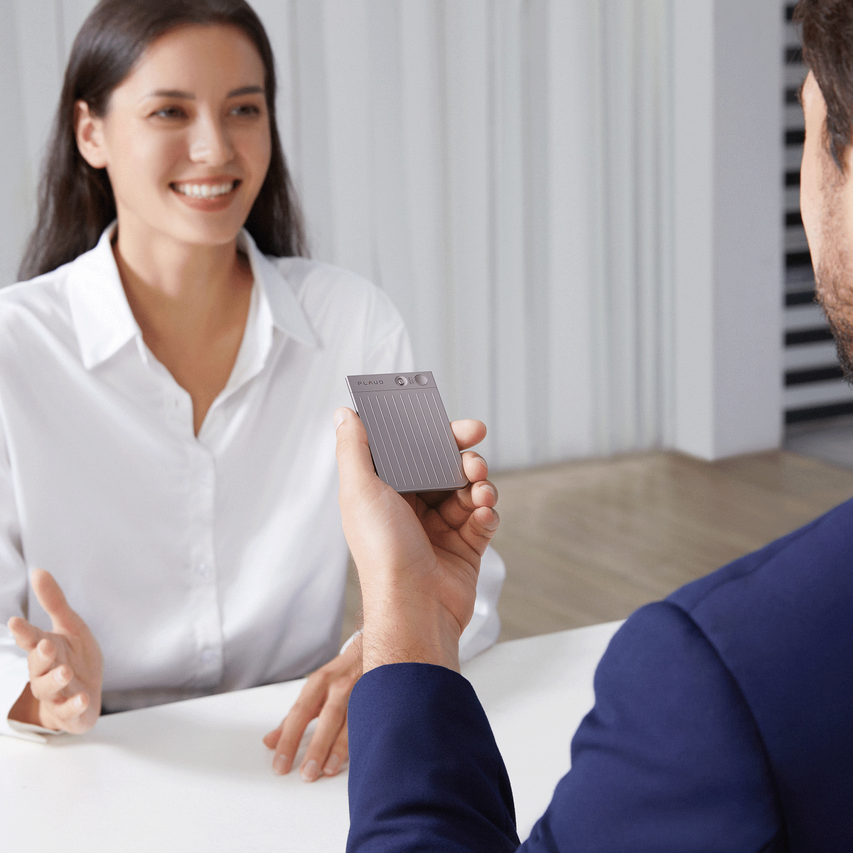 A professional holds a Plaud Note AI voice recorder during a conversation with a colleague in an office.