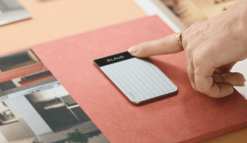 A finger presses the top of a silver Plaud NOTE AI voice recorder resting on a red folder.