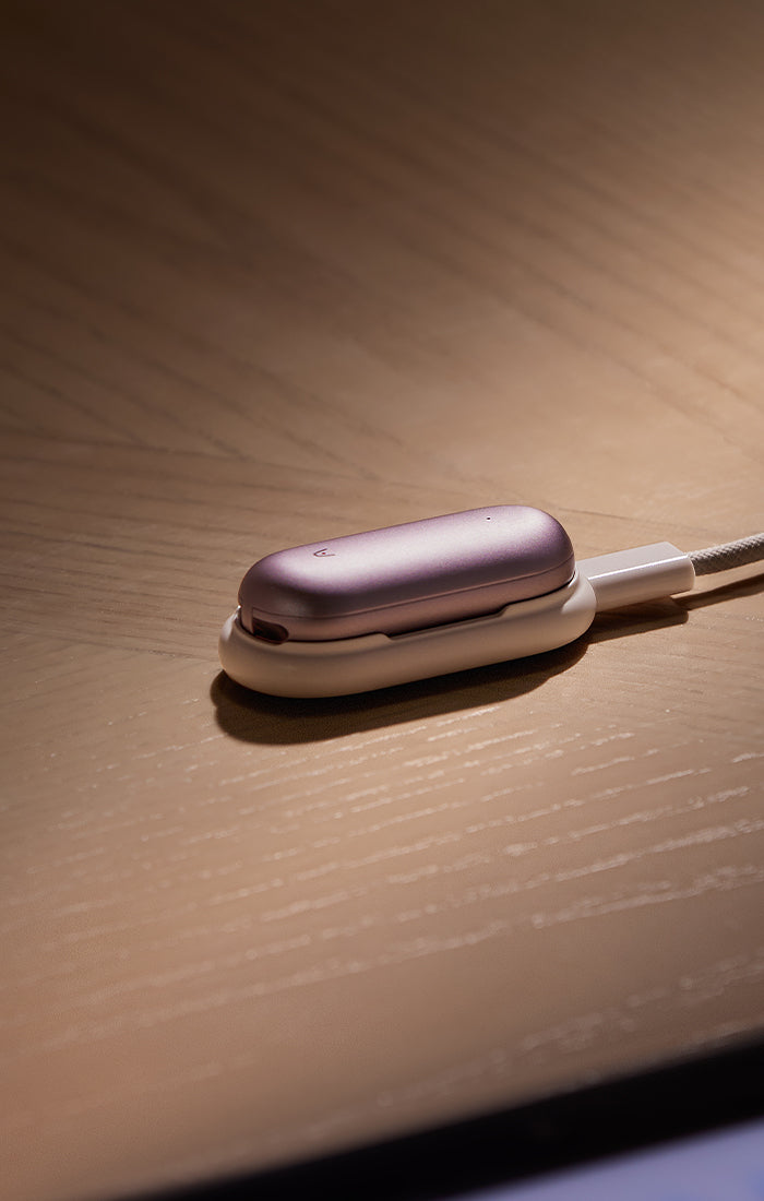 Rose gold Plaud NotePin AI voice recorder resting in its charging dock on a wooden surface.