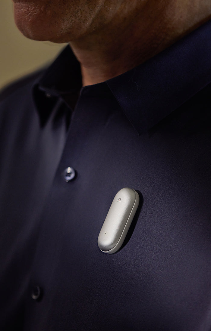 Silver PLAUD NotePin AI voice recorder clipped to a person's dark navy button-down shirt.