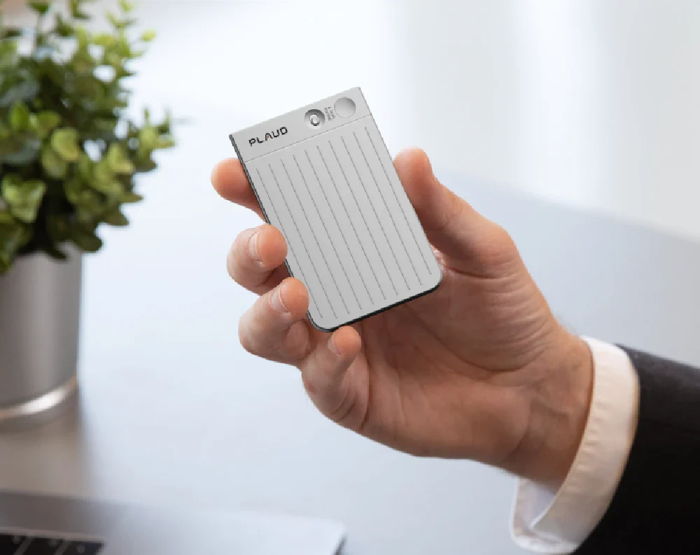 Hand holding the slim, silver PLAUD NOTE AI voice recorder in a professional office setting.