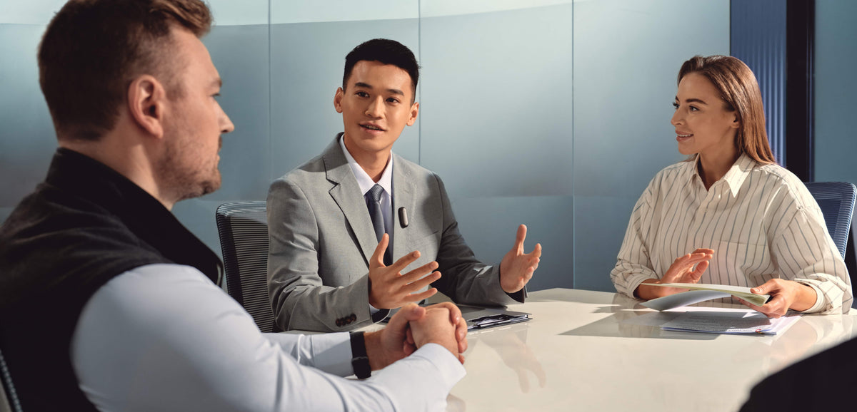 A professional man wearing a Plaud NotePin on his lapel during a business meeting with two colleagues.