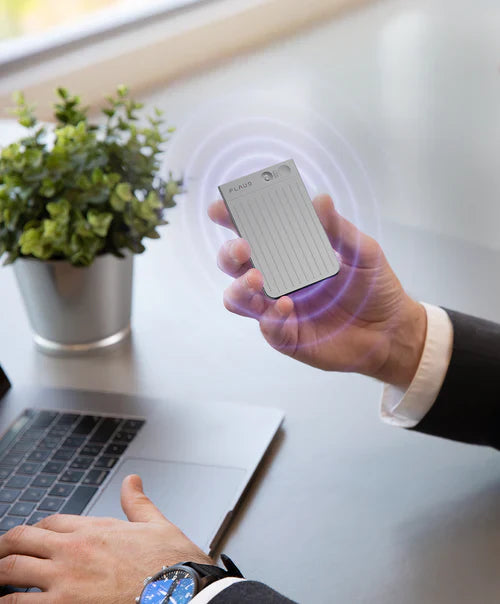Hand holding a silver Plaud Note AI voice recorder with purple sound wave graphics above a laptop.