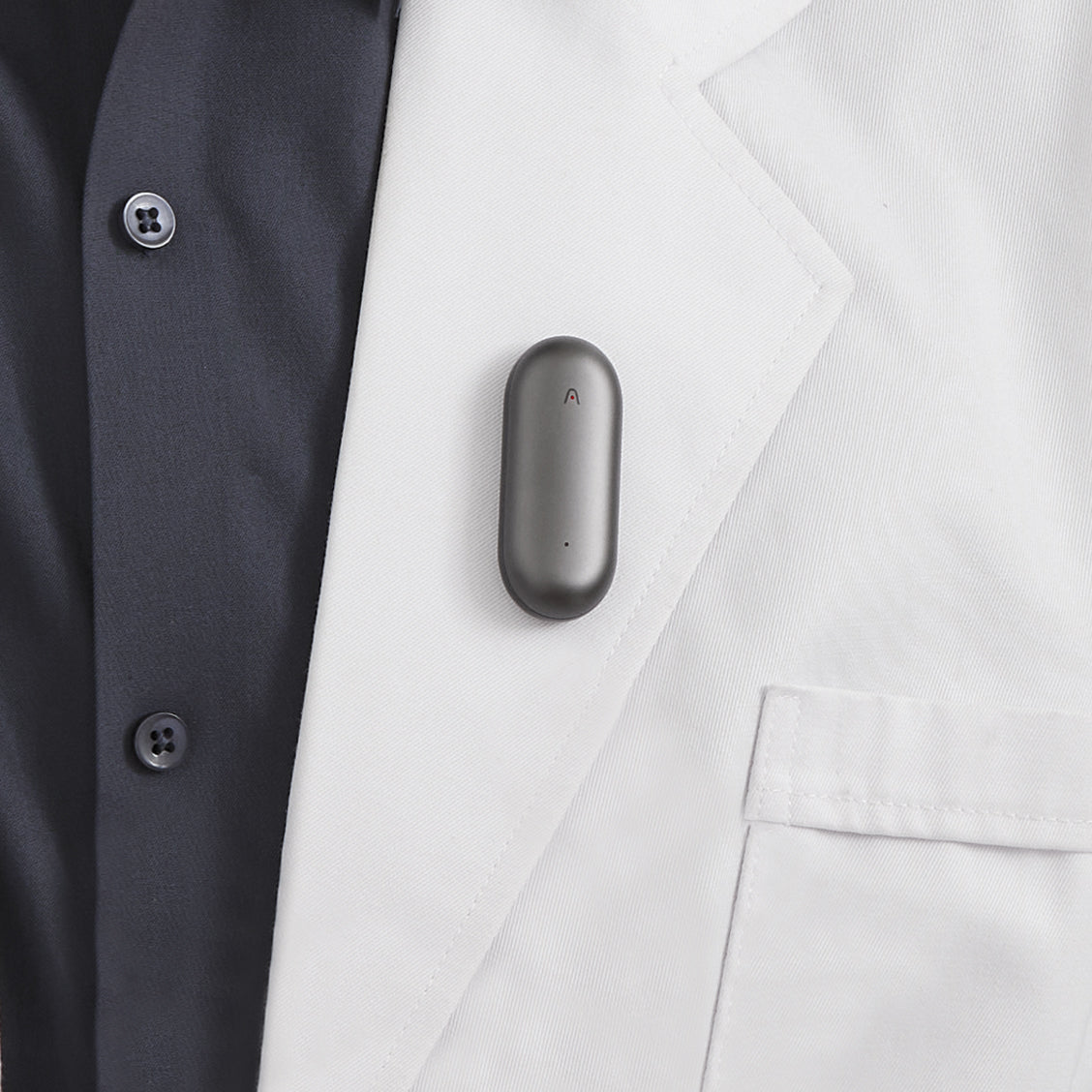 A gray Plaud NotePin AI voice recorder clipped to the lapel of a white lab coat.
