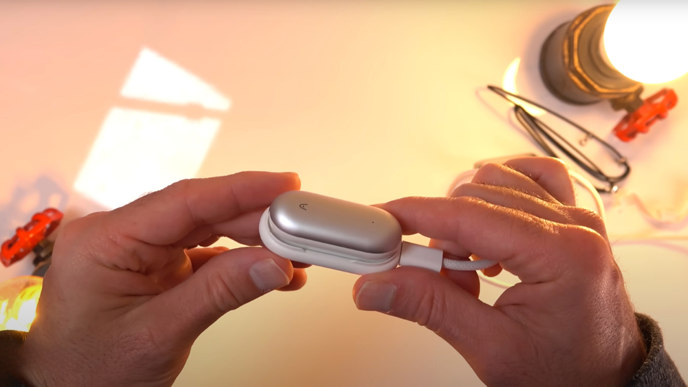 Hands holding the silver PLAUD NotePin AI voice recorder while it is connected to a white charging cable.