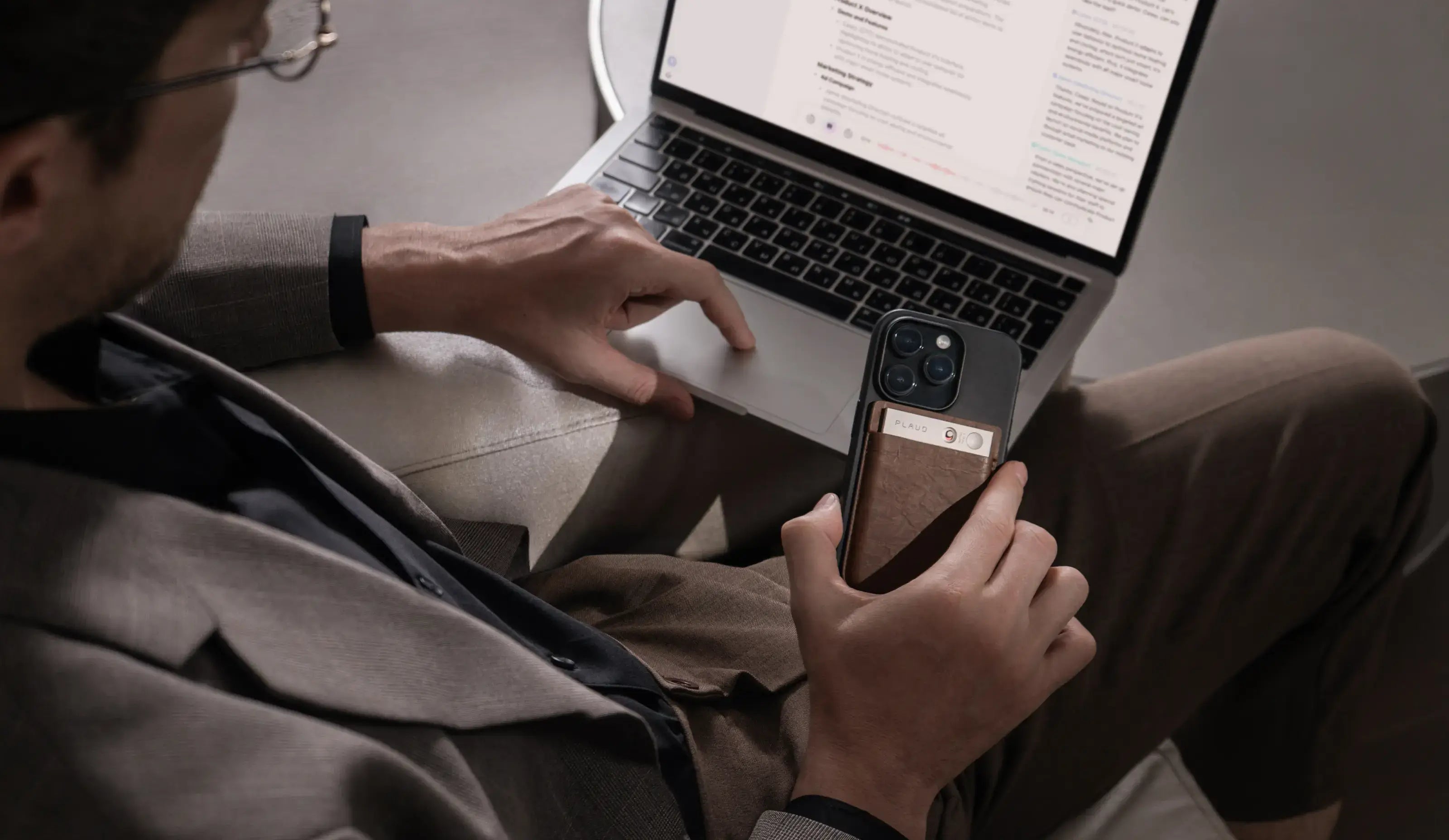 A professional using a laptop while holding a phone with a brown PLAUD NOTE AI voice recorder attached to the back.