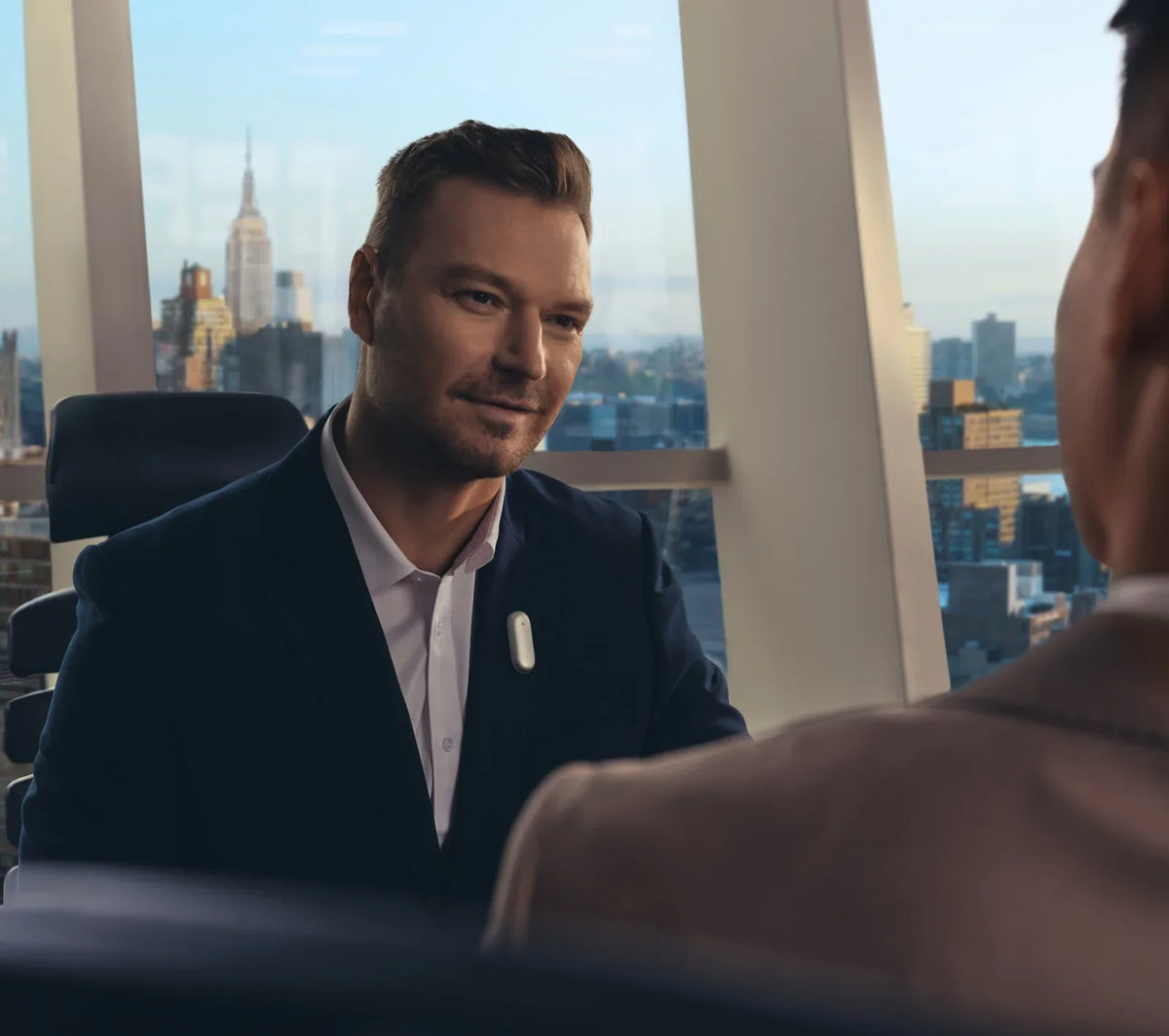 A businessman wearing a Plaud NotePin on his lapel during a meeting in a high-rise office with a New York City view.