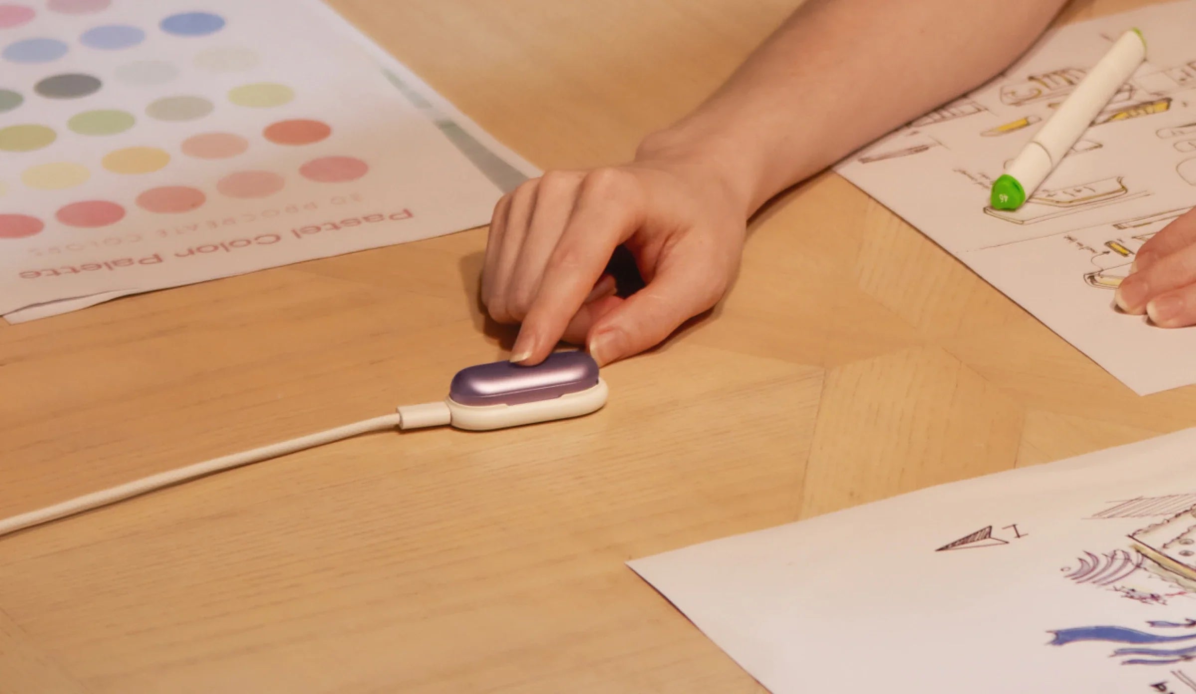 A hand presses the button on a purple PLAUD NotePin while charging on a desk surrounded by design sketches.