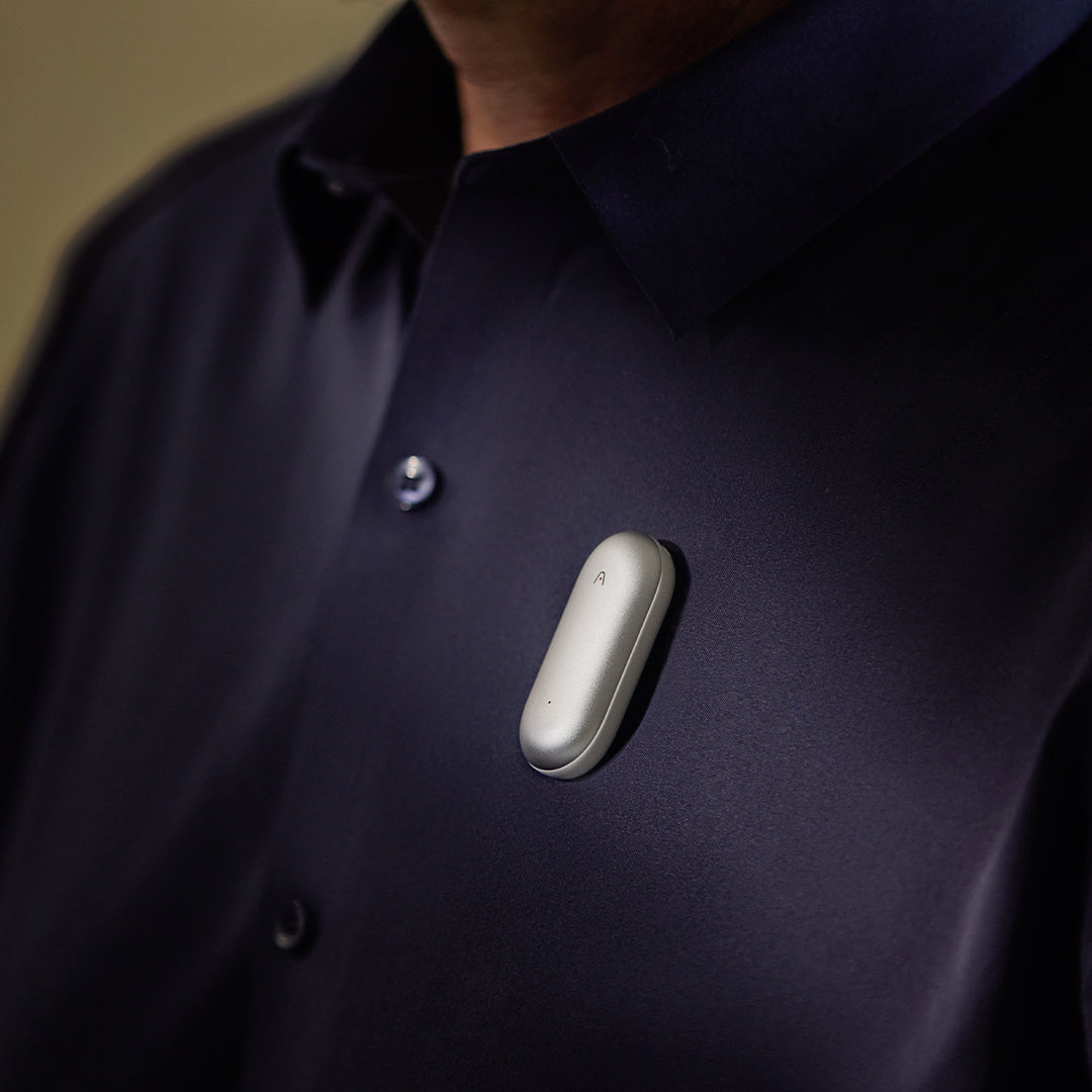 A silver Plaud NotePin AI voice recorder clipped onto a person's dark navy button-down shirt.
